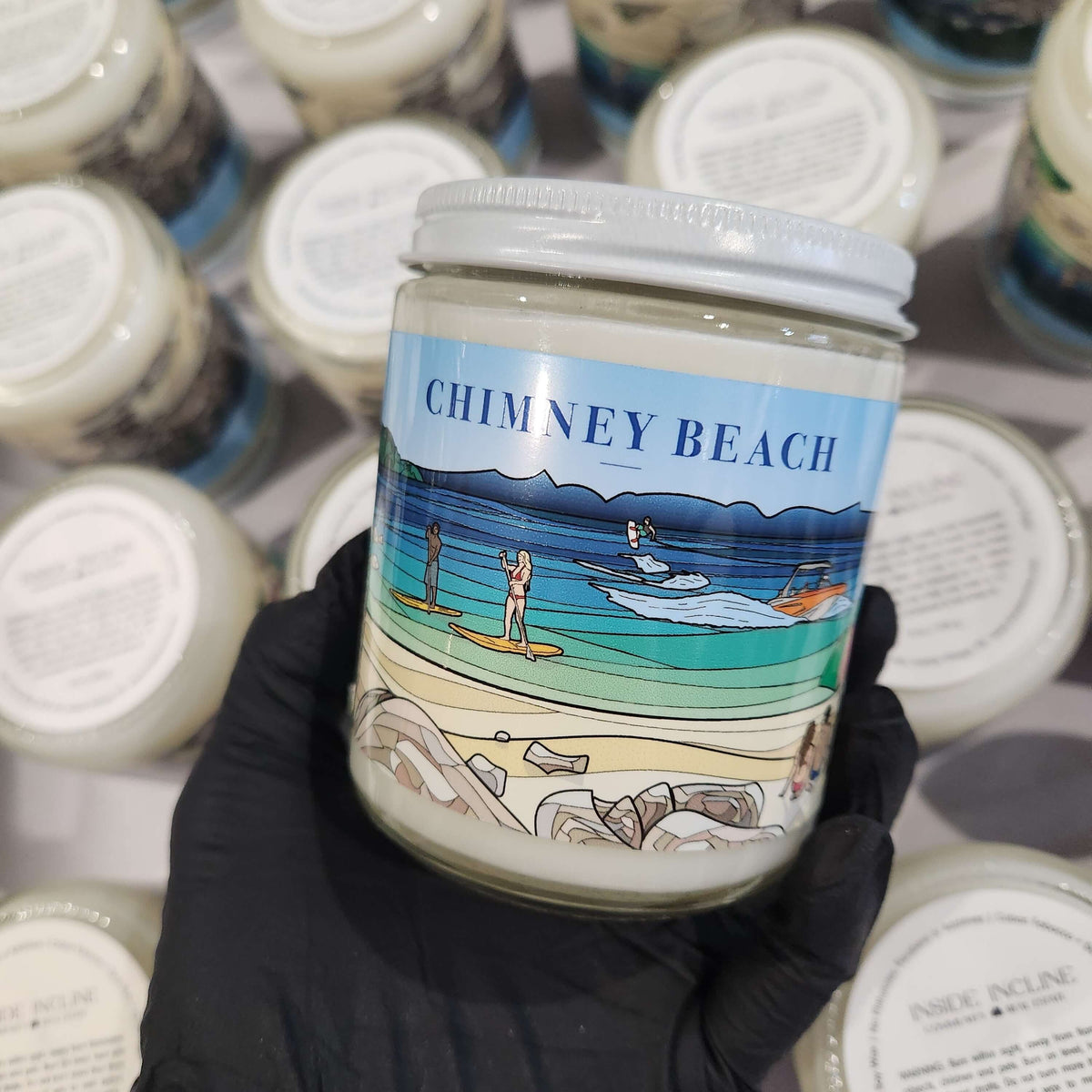 Candle jar with 'Chimney Beach' label held by a gloved hand, surrounded by similar jars.